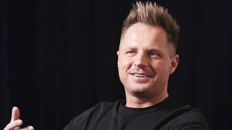 Photo of David Reger sitting on a couch on a stage, and gesturing with his hand while sharing an idea.