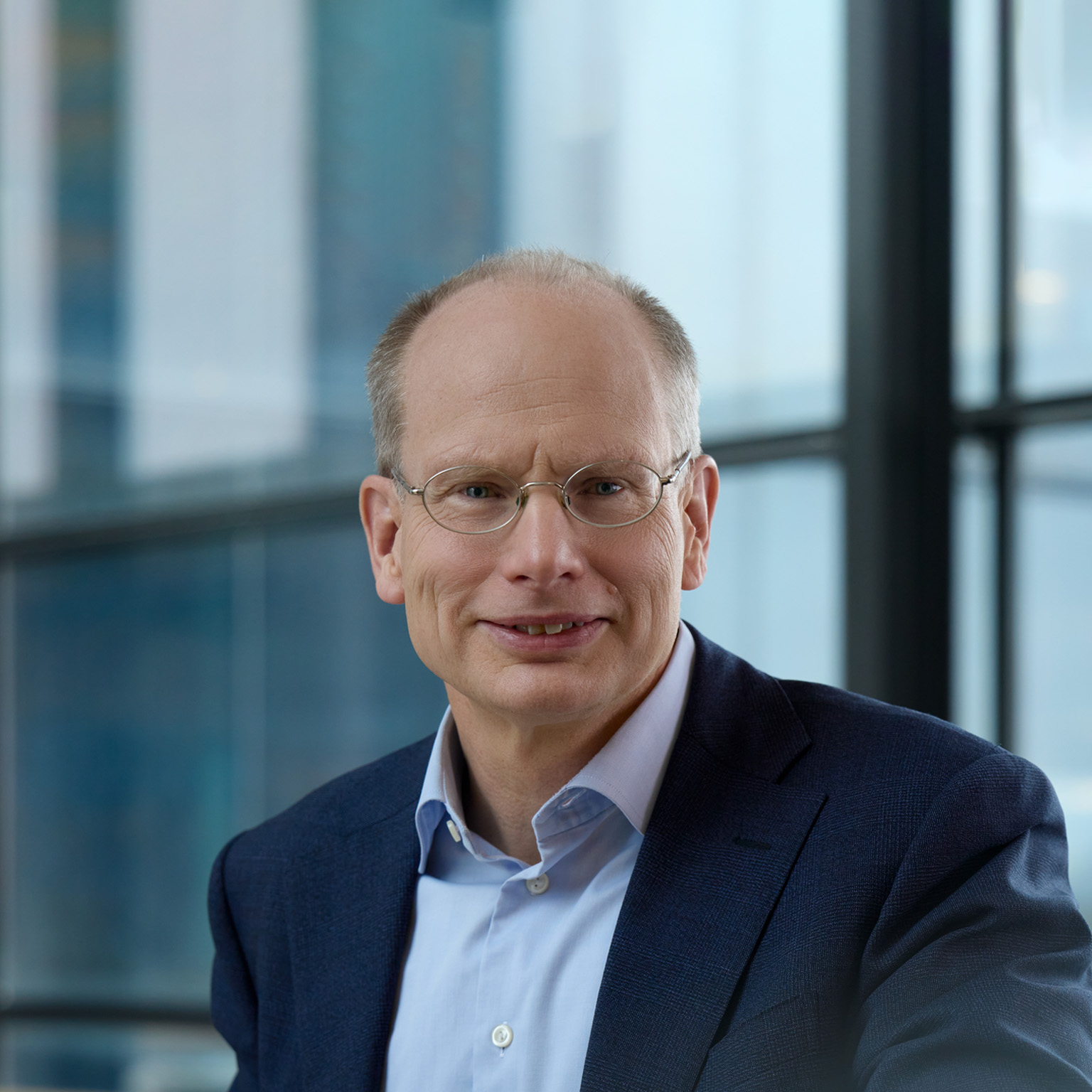 Professional portrait of Wärtsilä CEO Håkan Agnevall wearing thin-framed glasses, a navy blazer, and a light blue collared shirt, smiling slightly at the camera in front of large windows with a softly blurred blue background.