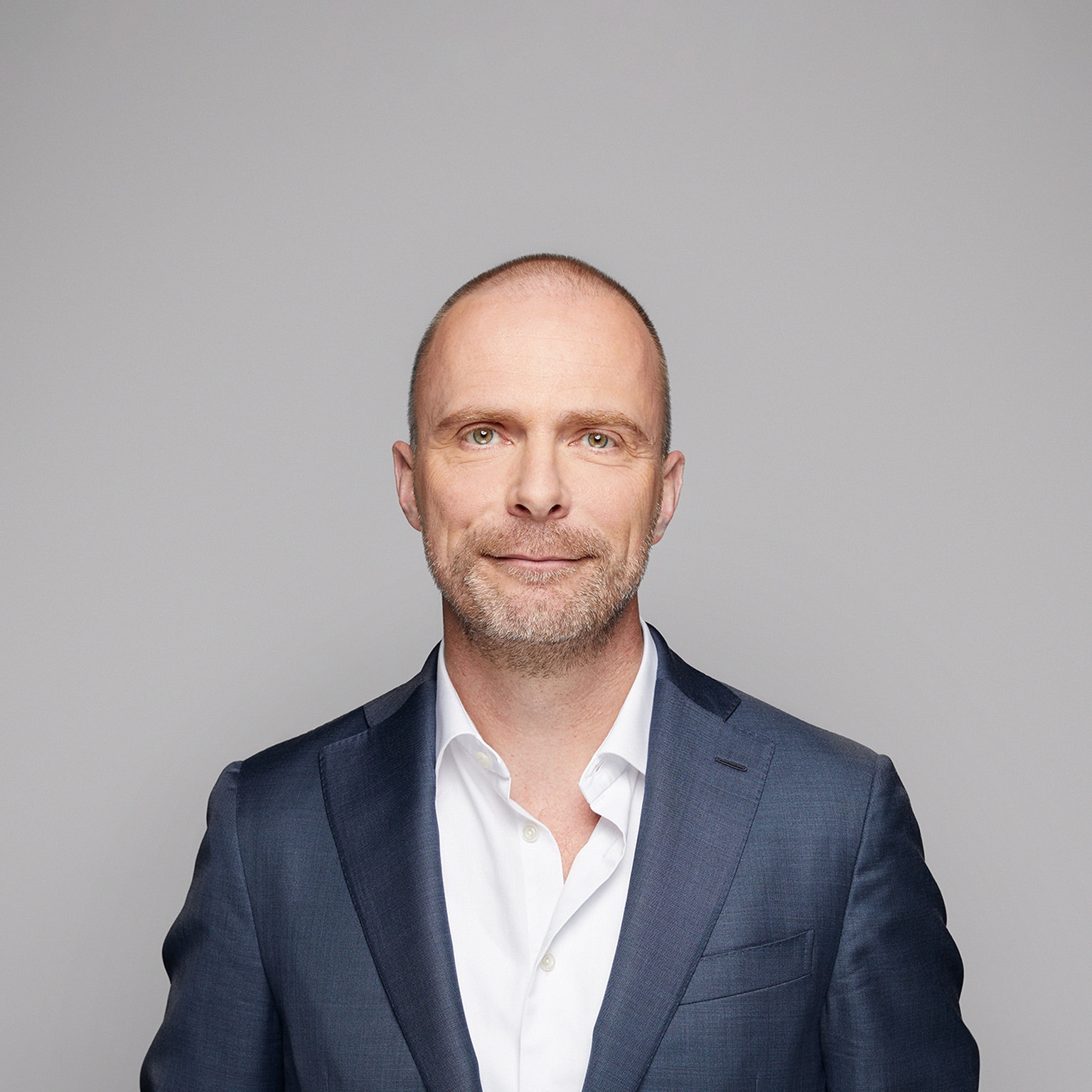 A portrait of Robert Carsouw, with a light beard, and a easy smile. He is wearing a dark blue suit and a white, open-collared shirt against a light gray background.