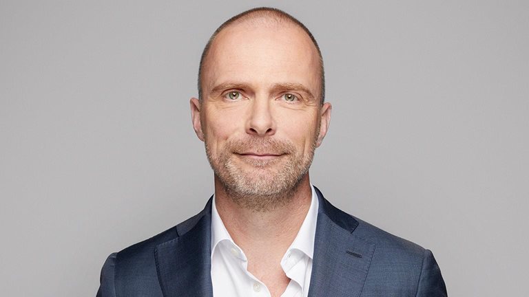 A portrait of Robert Carsouw, with a light beard, and a easy smile. He is wearing a dark blue suit and a white, open-collared shirt against a light gray background.