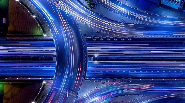 Aerial night view of a highway interchange with vibrant blue-toned long-exposure light trails from moving traffic.