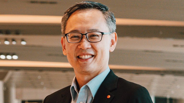 Lim Ching Kiat smiles while standing in an airport, dressed in a suit and wearing glasses.