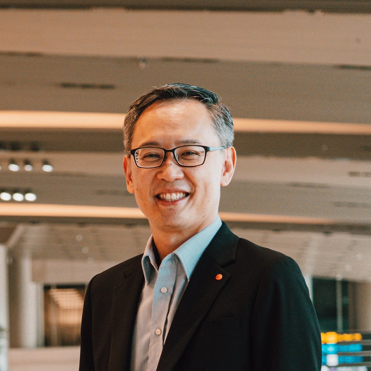 Lim Ching Kiat smiles while standing in an airport, dressed in a suit and wearing glasses.