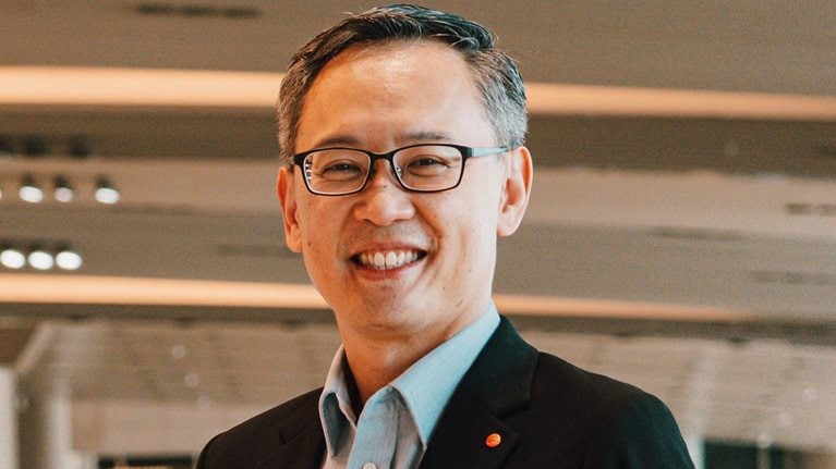 Lim Ching Kiat smiles while standing in an airport, dressed in a suit and wearing glasses.