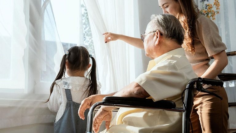 Elderly man in a wheelchair looks out a window with granddaughter and care giver