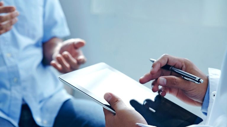 A close up of a doctor's hands holding a tablet while talking to a patient about his treatment plan.
