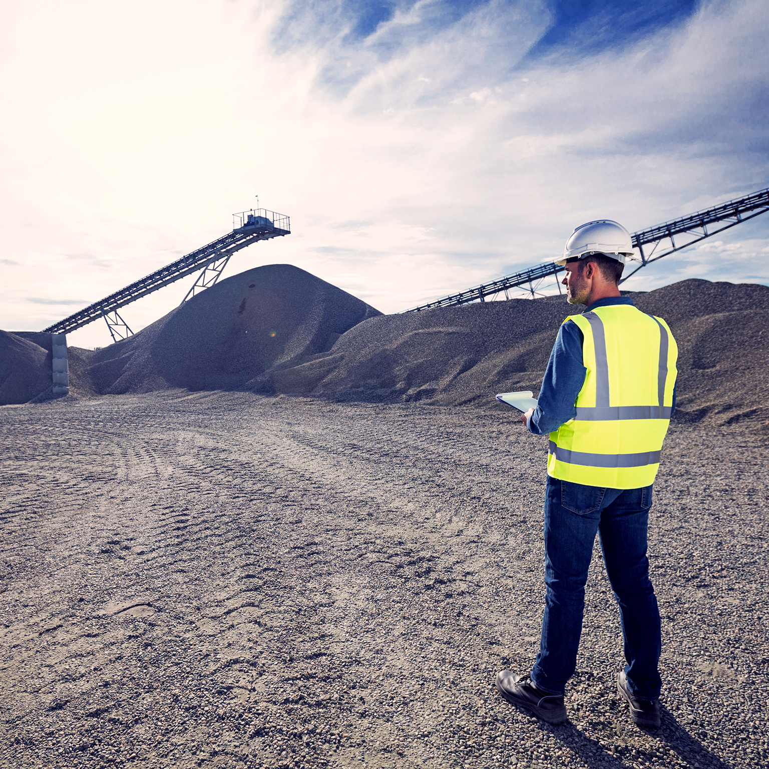 Open-pit mine engineer wearing protective clothes and white helmet standing in front of pile of gravel, taking notes on clipboard. Quality control concept.