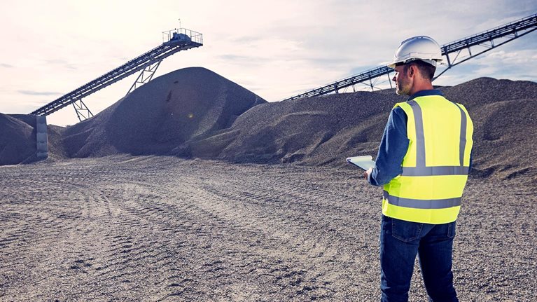 Open-pit mine engineer wearing protective clothes and white helmet standing in front of pile of gravel, taking notes on clipboard. Quality control concept.
