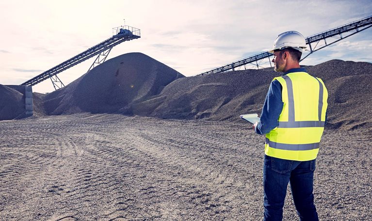 Open-pit mine engineer wearing protective clothes and white helmet standing in front of pile of gravel, taking notes on clipboard. Quality control concept.