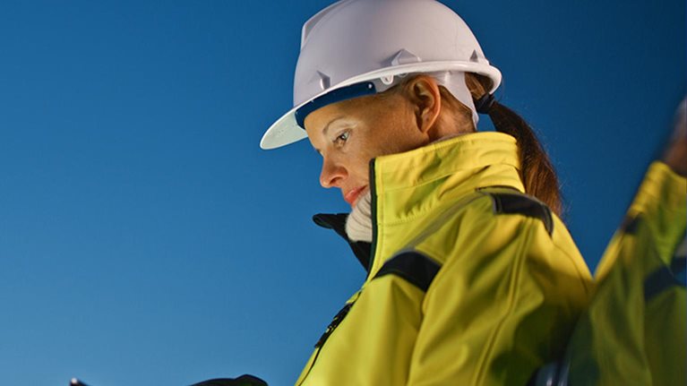 Female worker in protective workwear using tablet PC while standing by car at oil industry against sky