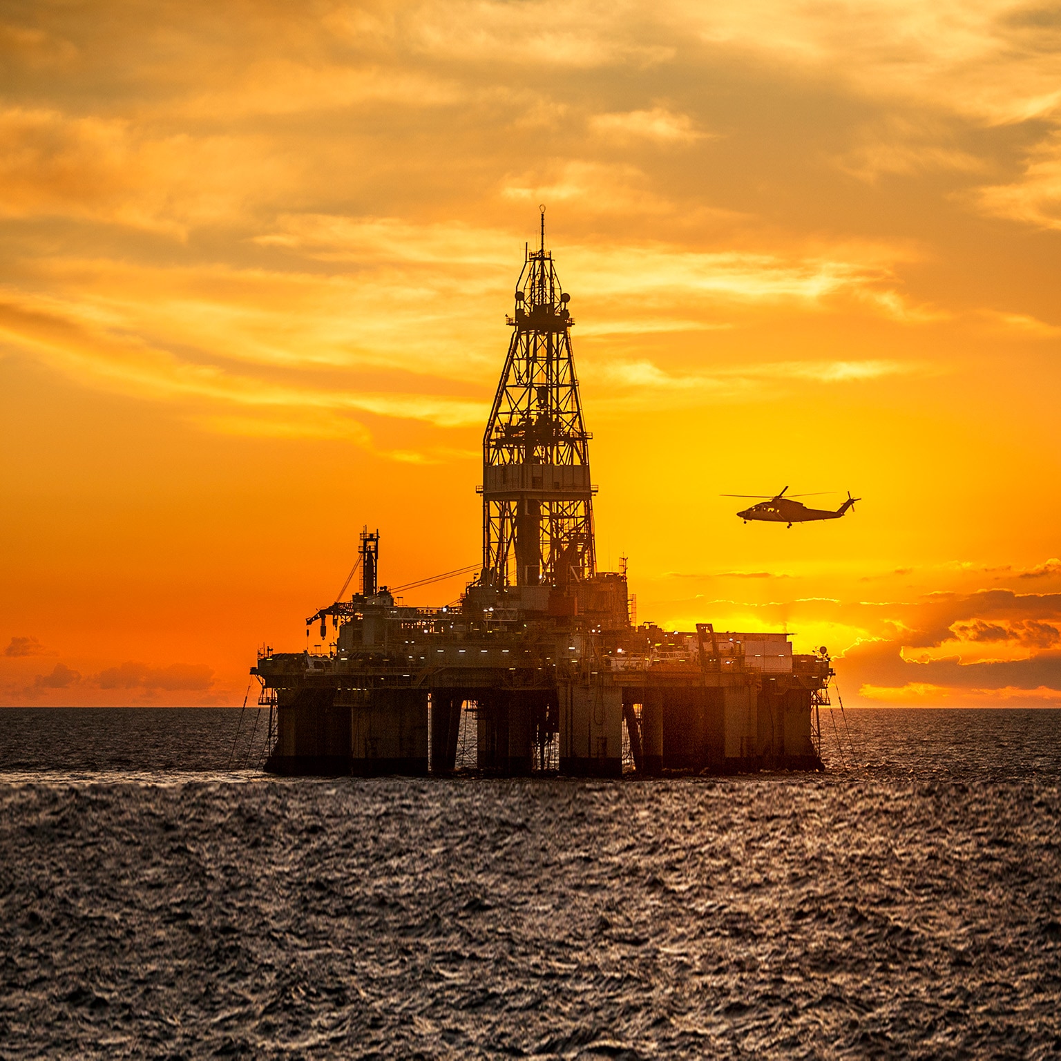 Helicopter flying over oil rig in sea against sky during sunset - stock photo