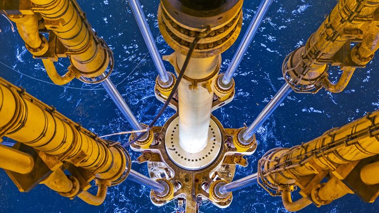 Looking down, we can see an oil rig's drill in the North Sea. The rig's equipment is painted a vibrant shade of yellow and we can see the deep blue water and waves beneath it.