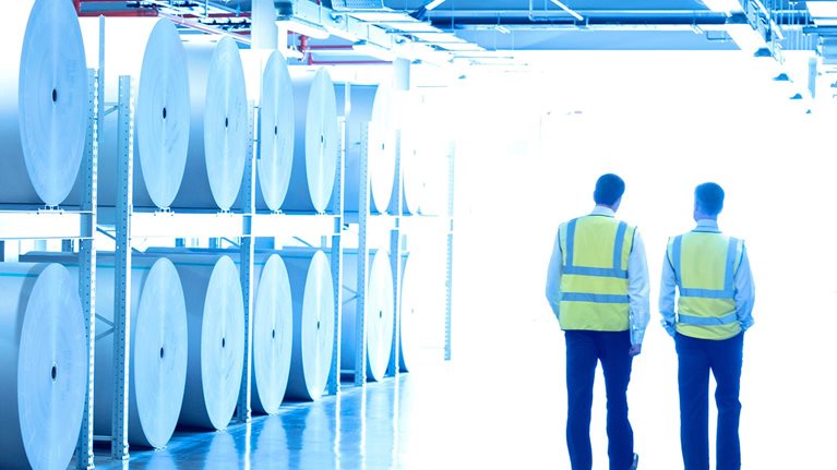 Workers in reflective clothing walking along large paper spools in printing plant