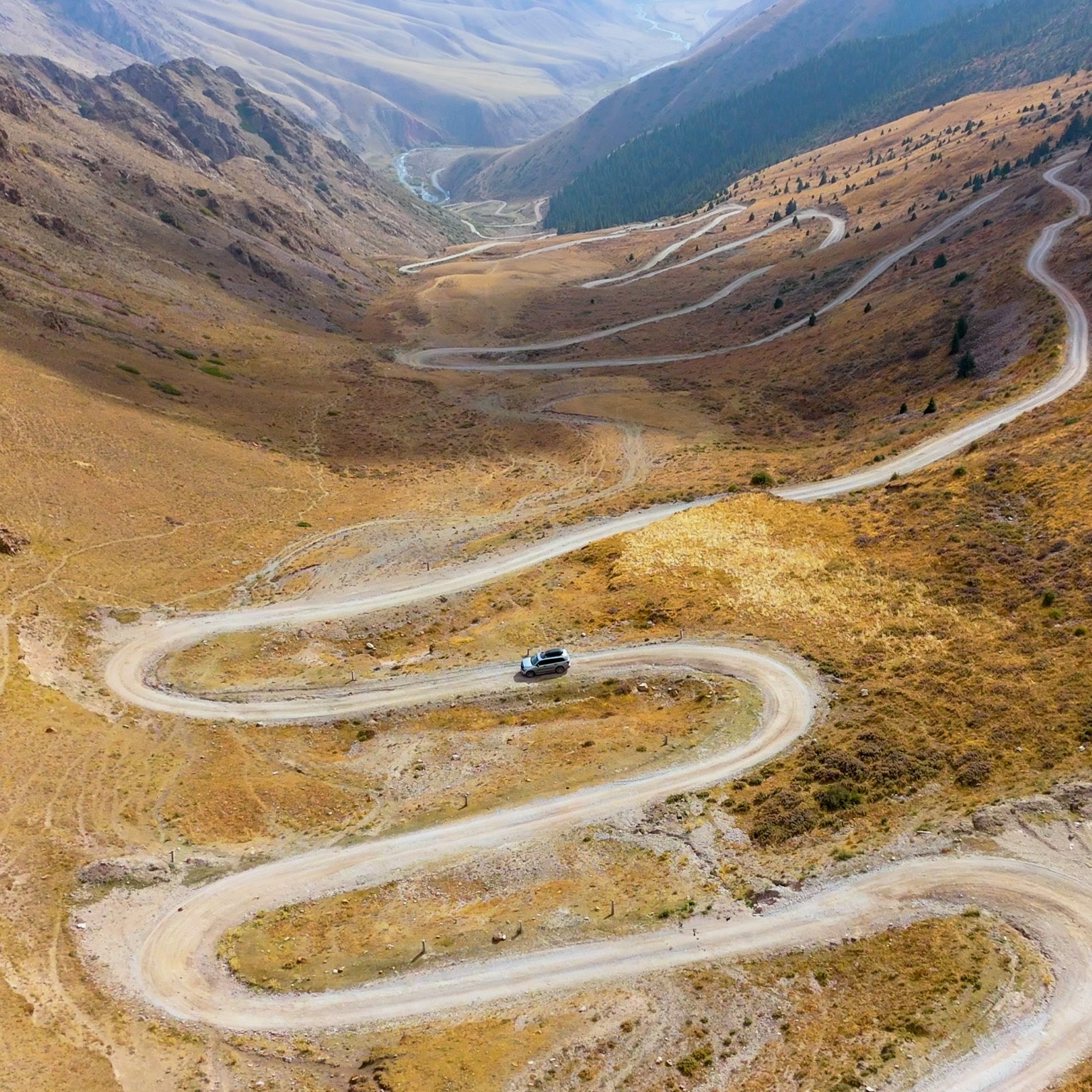 A silver SUV makes navigates a winding dirt road as it snakes through a wide mountain valley, cutting sharp switchbacks across dry, golden-brown slopes. Steep ridges rise on both sides, with sparse vegetation and distant peaks fading into a hazy, expansive backdrop.