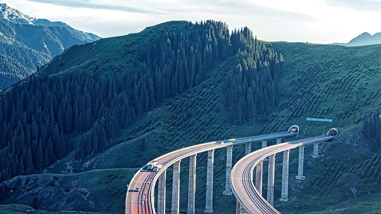 A winding elevated highway curves gracefully through a lush green mountainous landscape, supported by tall pillars. In the distance, forested hills and rugged peaks stretch beneath a bright, partly cloudy sky.