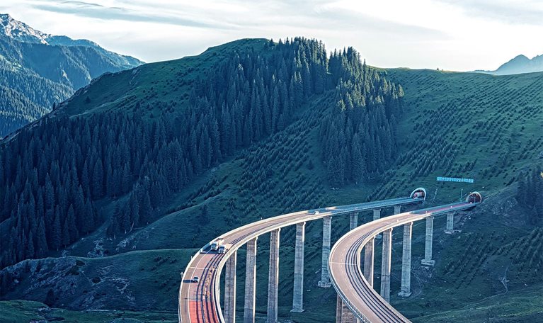 A winding elevated highway curves gracefully through a lush green mountainous landscape, supported by tall pillars. In the distance, forested hills and rugged peaks stretch beneath a bright, partly cloudy sky.