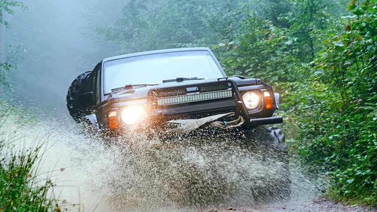 The image shows a rugged off-road vehicle driving through a muddy forest trail, splashing water as it moves forward. Dense green foliage surrounds the path, and mist hangs in the air, creating a dramatic and adventurous atmosphere.
