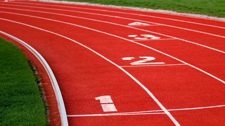 An empty red running track curves through a green field, with white lane markings and the numbers one through four visible, suggesting competition, positioning, or the importance of starting point.