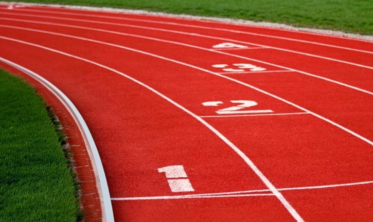 An empty red running track curves through a green field, with white lane markings and the numbers one through four visible, suggesting competition, positioning, or the importance of starting point.