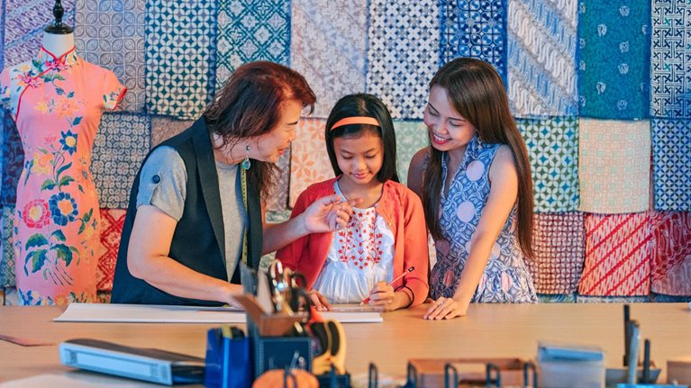 A Chinese grandmother teaching her granddaughter how to design a garment while her mother looks on. Fabric patterns hang on the wall behind them and traditional Chinese dresses hang on mannequins.  
