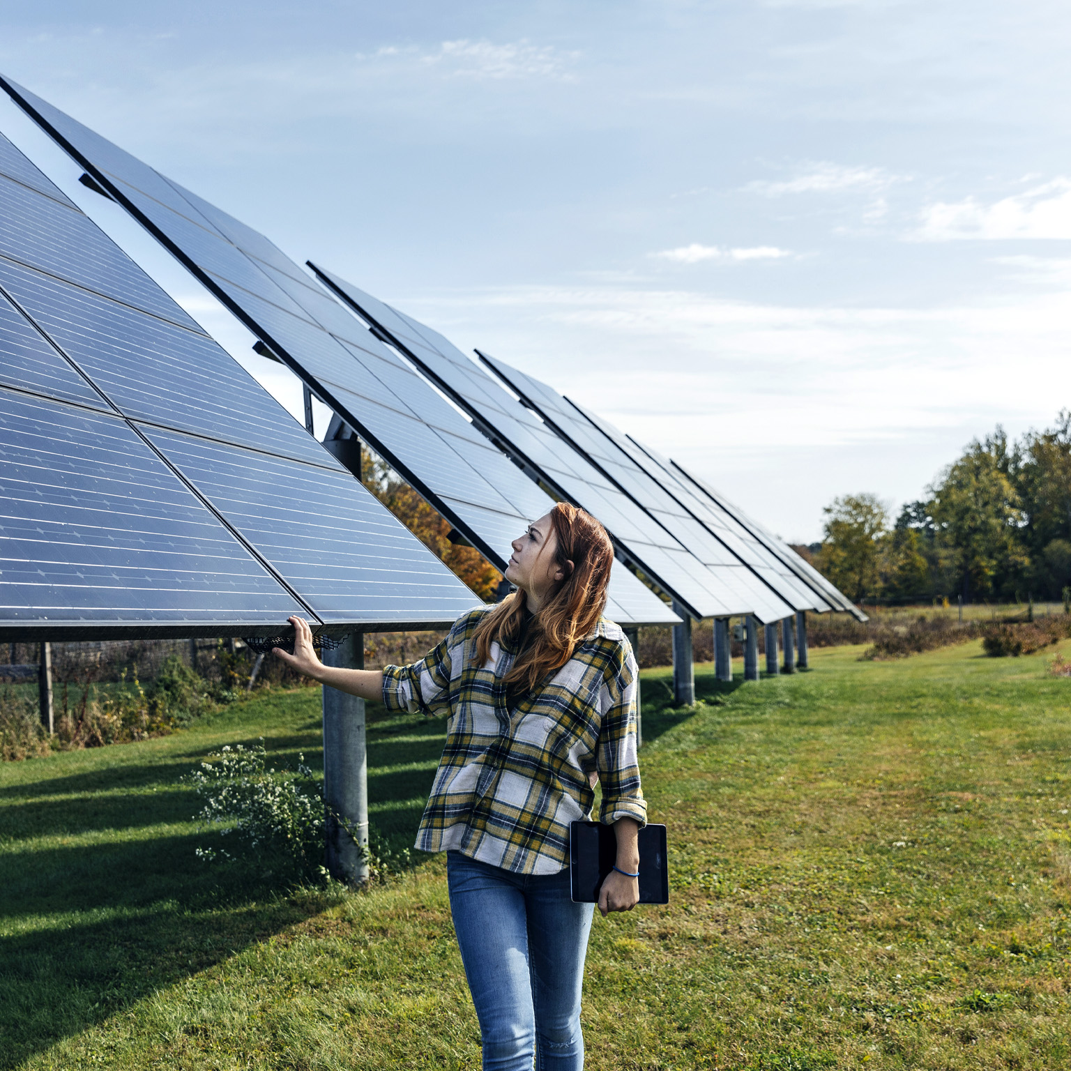 A young woman in a flannel shirt inspects a row of solar panels on a farm in upstate New York. She carefully places her hand on one of them. 