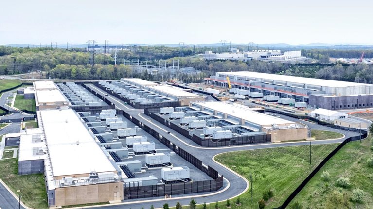 A sprawling data center complex surrounded by greenery and infrastructure. Several rectangular buildings with white roofs are visible, densely packed with cooling units or similar equipment, with at least one building under construction.