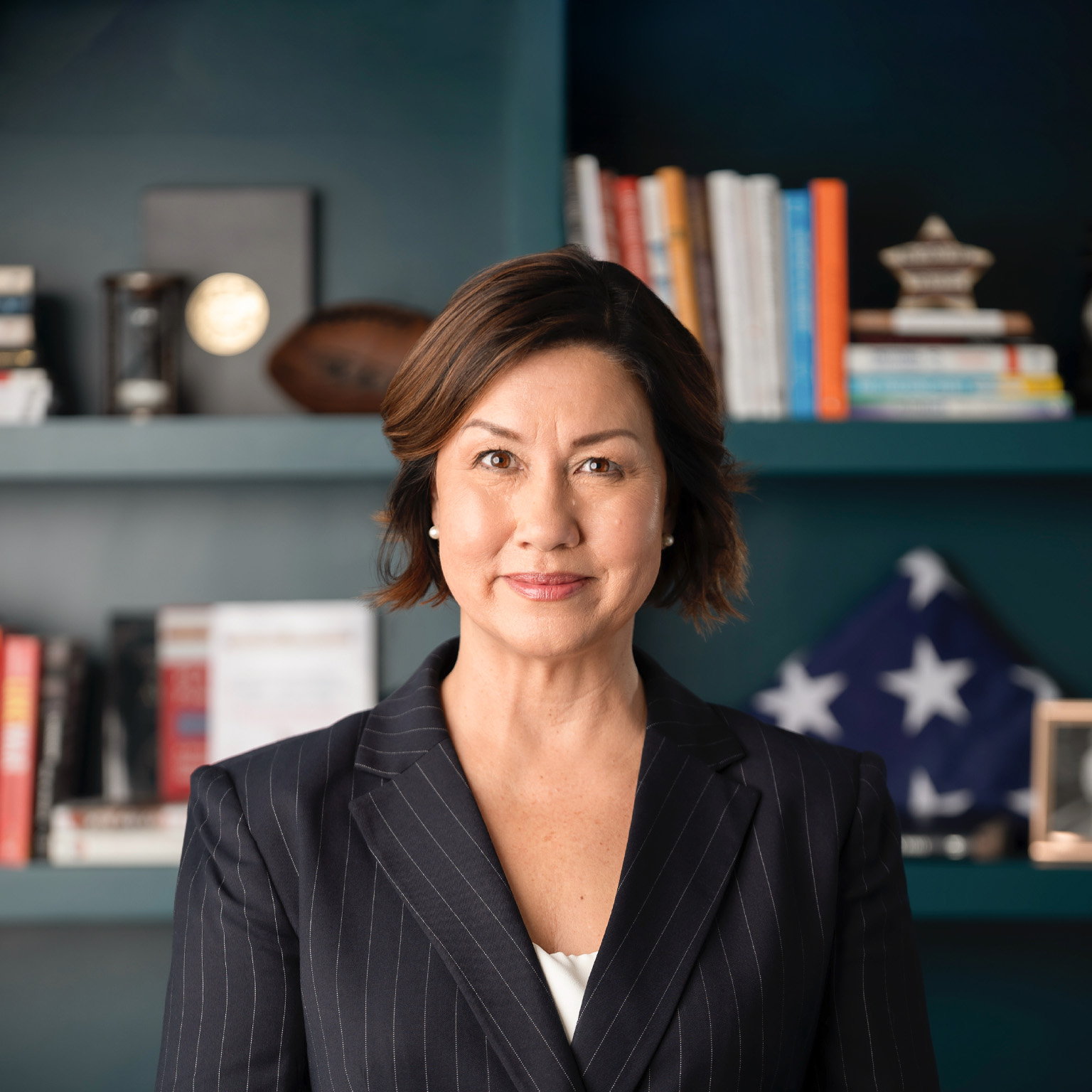 A professional portrait of JoAnne Bass in a dark pinstripe blazer, facing the camera with a composed expression. Behind her, softly blurred shelves with books, decorative objects, and a folded American flag.