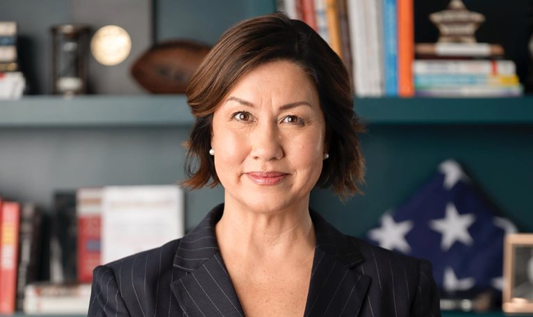 A professional portrait of JoAnne Bass in a dark pinstripe blazer, facing the camera with a composed expression. Behind her, softly blurred shelves with books, decorative objects, and a folded American flag.
