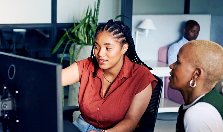 Two women collaborating closely at a computer, intensely focused on the screen. The one woman gestures towards the monitor, suggesting an learning or problem-solving session.