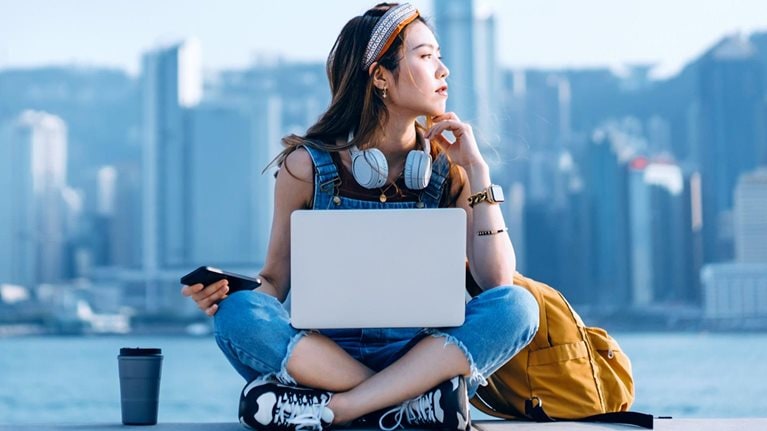 A young person, with a backpack, a beverage tumbler, and a laptop, is sitting on a ledge with back to the waterbody against the backdrop of skyscrapers.