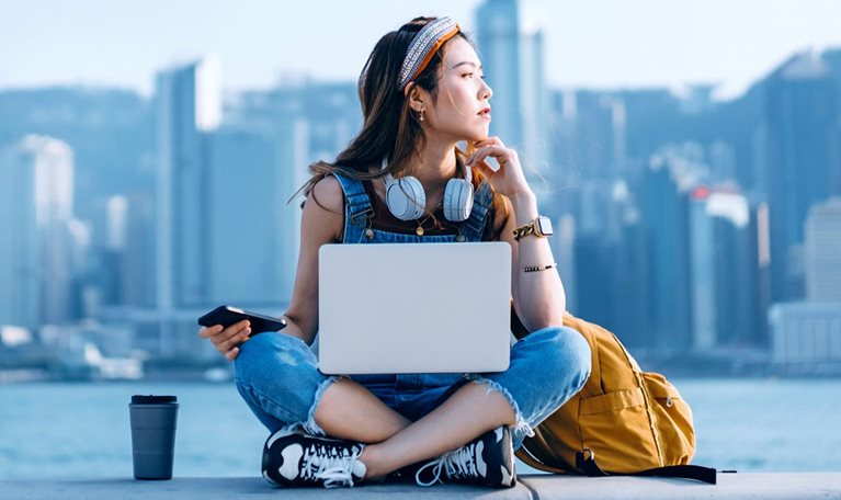 A young person, with a backpack, a beverage tumbler, and a laptop, is sitting on a ledge with back to the waterbody against the backdrop of skyscrapers.