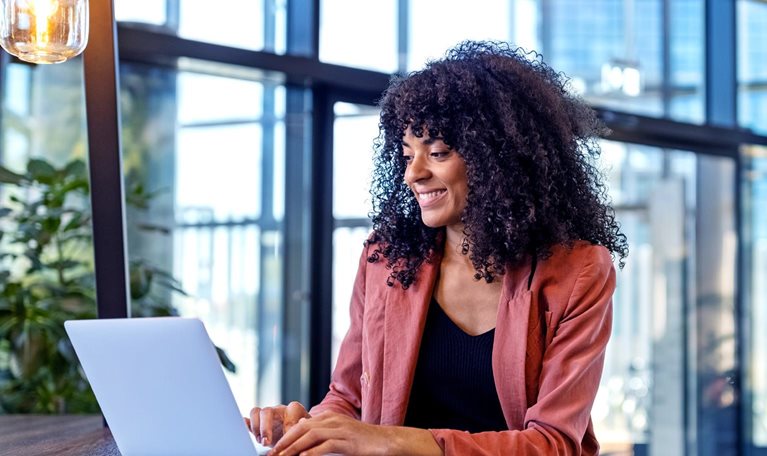 A cheerful young lady is seated at a table in a contemporary and well-lit cafe, typing away on her laptop.