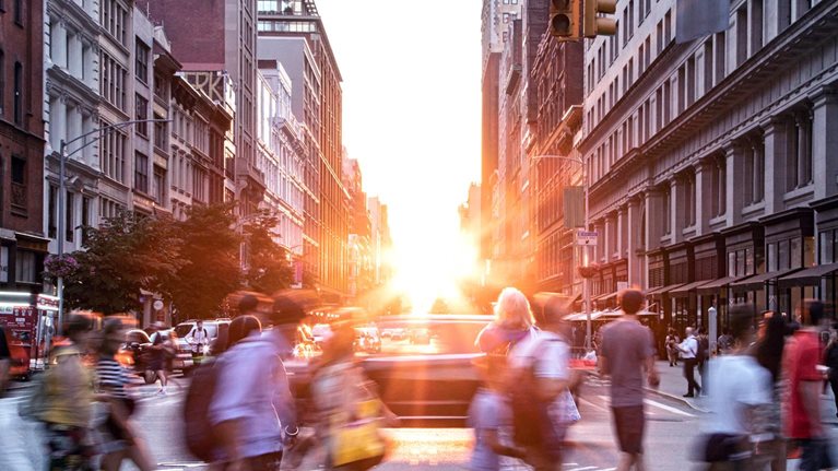 A diverse crowd of people walking through a bustling downtown intersection with the sunset in the background.