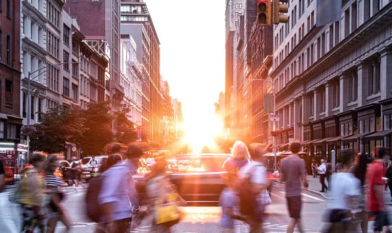 A diverse crowd of people walking through a bustling downtown intersection with the sunset in the background.