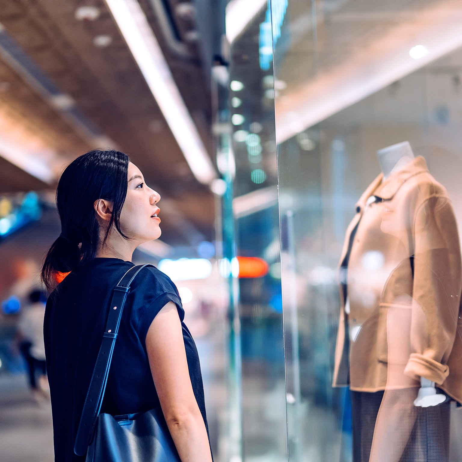 Young Asian woman standing outside a boutique looking at window display while shopping in shopping mall. Window shopping. Sale season