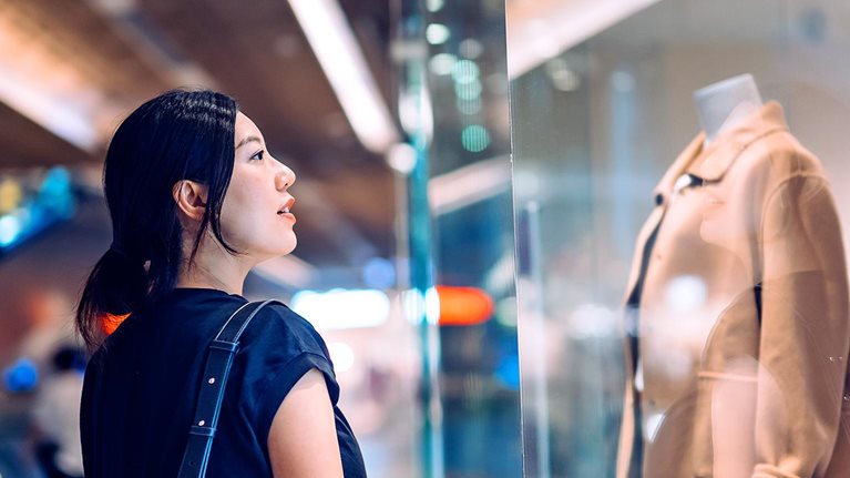 Young Asian woman standing outside a boutique looking at window display while shopping in shopping mall. Window shopping. Sale season