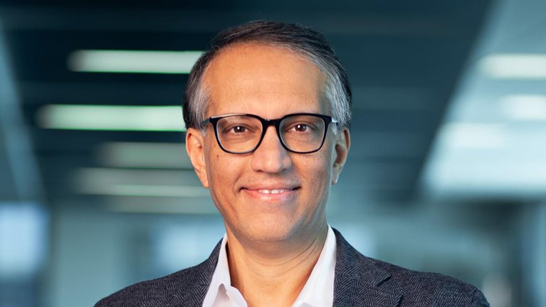 Portrait of a smiling Vipul Chawla shot against a blurred out office setting. Chawla is wearing glasses and a dark gray sport coat and button down shirt.