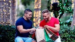 Two men dressed for warmer weather look through their purchases while holding holiday themed shopping bags. String lights adorn the trees behind them.