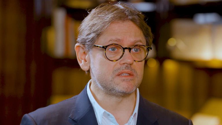 Laurent Kleitman wearing round glasses, a navy blazer, and a light shirt, speaking to Alex Sawaya during an interview. 