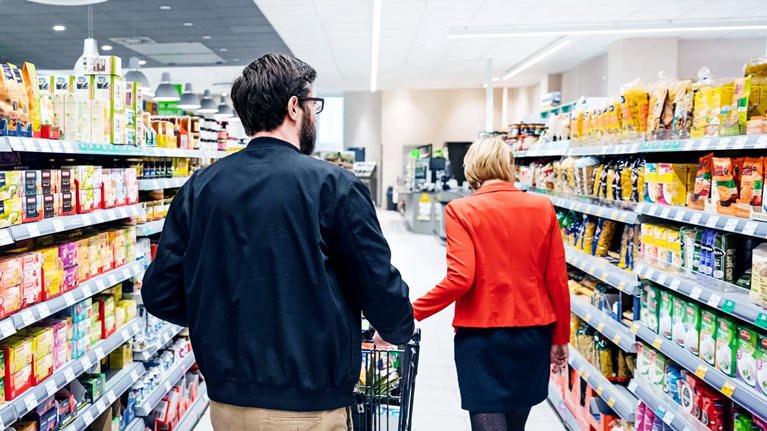 A couple seen from behind, walking down an aisle stocked with boxed goods at their local supermarket
