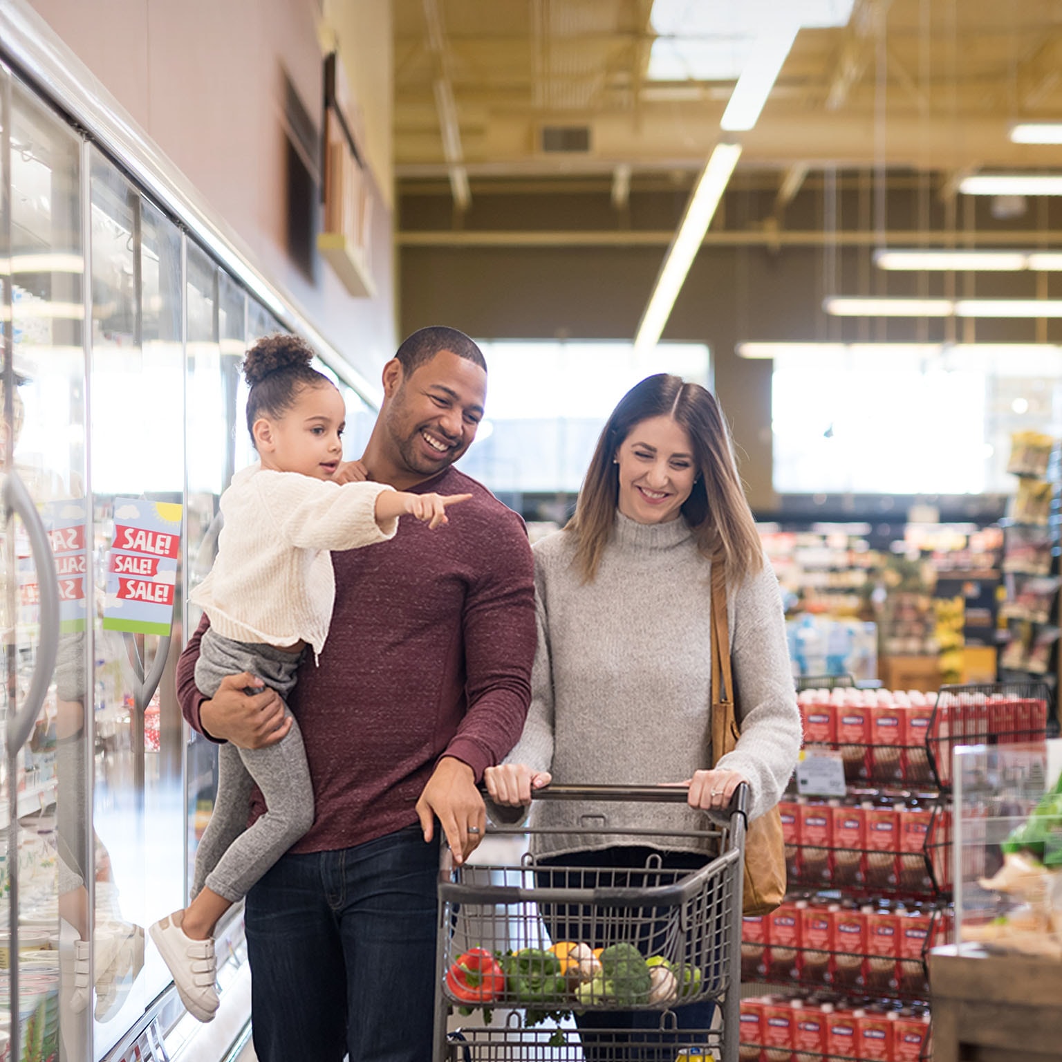 Dad holds his pre-school age daughter at the grocery. Mom is next to him pushing the cart. They are both chuckling as they stroll through the refrigerated dairy section.