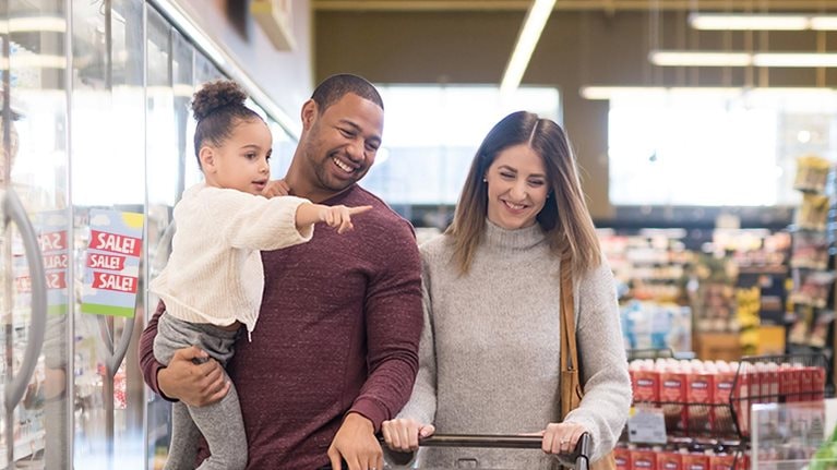 Dad holds his pre-school age daughter at the grocery. Mom is next to him pushing the cart. They are both chuckling as they stroll through the refrigerated dairy section.