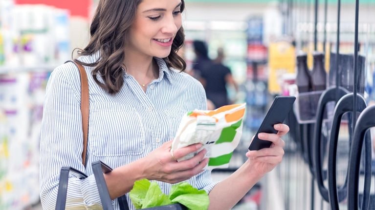 Confident young Hispanic woman holds smart phone as she reads a nutrition label on a bag of frozen vegetables. She is holding a shopping basket filled with healthy foods.