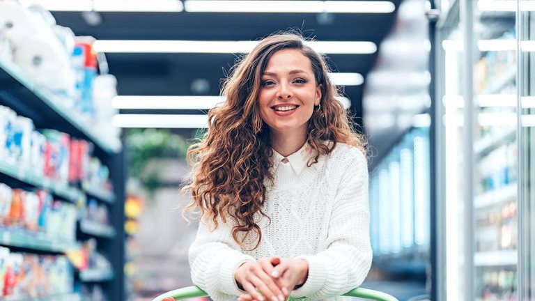 A young woman stands in a bright, well-organized grocery store, carefully selecting products from the shelves. Surrounded by neatly arranged goods and fresh selections, the scene captures everyday consumer behavior, independence, and healthy decision-making in a contemporary retail environment.
