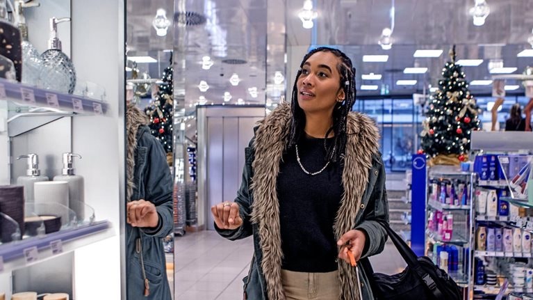 A woman walking down an aisle in a store while looking at personal care items. She is wearing a warm coat and holding a shopping basket, store holiday decorations can be seen in the background.