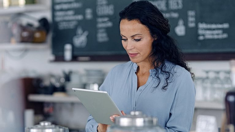 Brunette woman using digital tablet while standing at counter in coffee shop with the menu behind her.