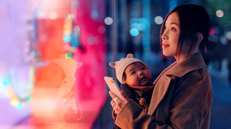 Mother carrying her baby and using a smartphone while looking at a brightly lit shop window display.