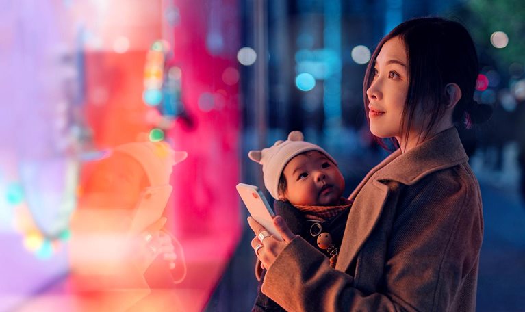 Mother carrying her baby and using a smartphone while looking at a brightly lit shop window display.