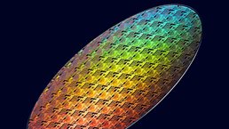 Colorful iridescent semiconductor wafer with intricate microchip patterns displayed against a dark background.
