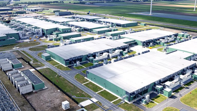 High angle view of a data center surrounded by green fields and wind turbines on the horizon.